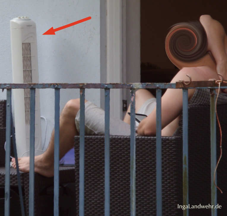 Ein Ventilator steht auf dem Balkon. Davor sitzt ein Mann und liest.