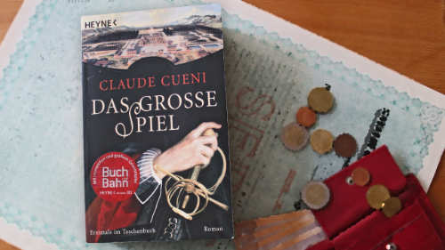 Neben dem Buch "Das große Spiel" von Claude Cueni liegen Geldscheine und Münzen. Alles liegt auf einer alten Aktie.