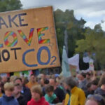 Menschen, die für Nachhaltigkeit demonstrieren. Im Vordergrund ein Schild mit der Aufschrift "Make Love not CO2"