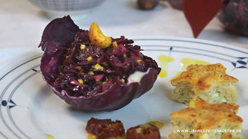 Rotkohlsalat ist in einem Rotkohlblatt angerichtet. Darauf liegt ein Cashewkern und daneben zwei Hüttenkäse-Küchlein