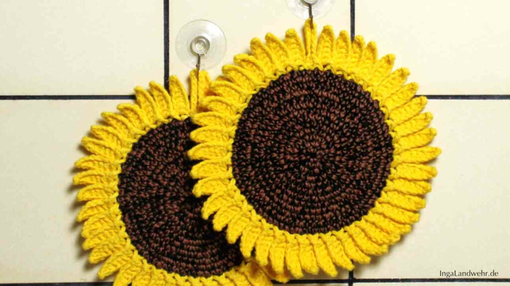 Zwei gehäkelte Sonnenblumen-Topflappen hängen an einer gefliesten Wand.