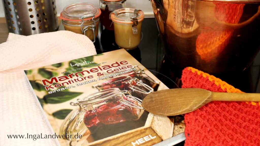 Neben dem Buch Marmelade, Konfitüre & Gelee steht ein Kupfertopf, davor liegt ein Kochlöffel und im Hintergrund stehen kleine Marmeladengläser